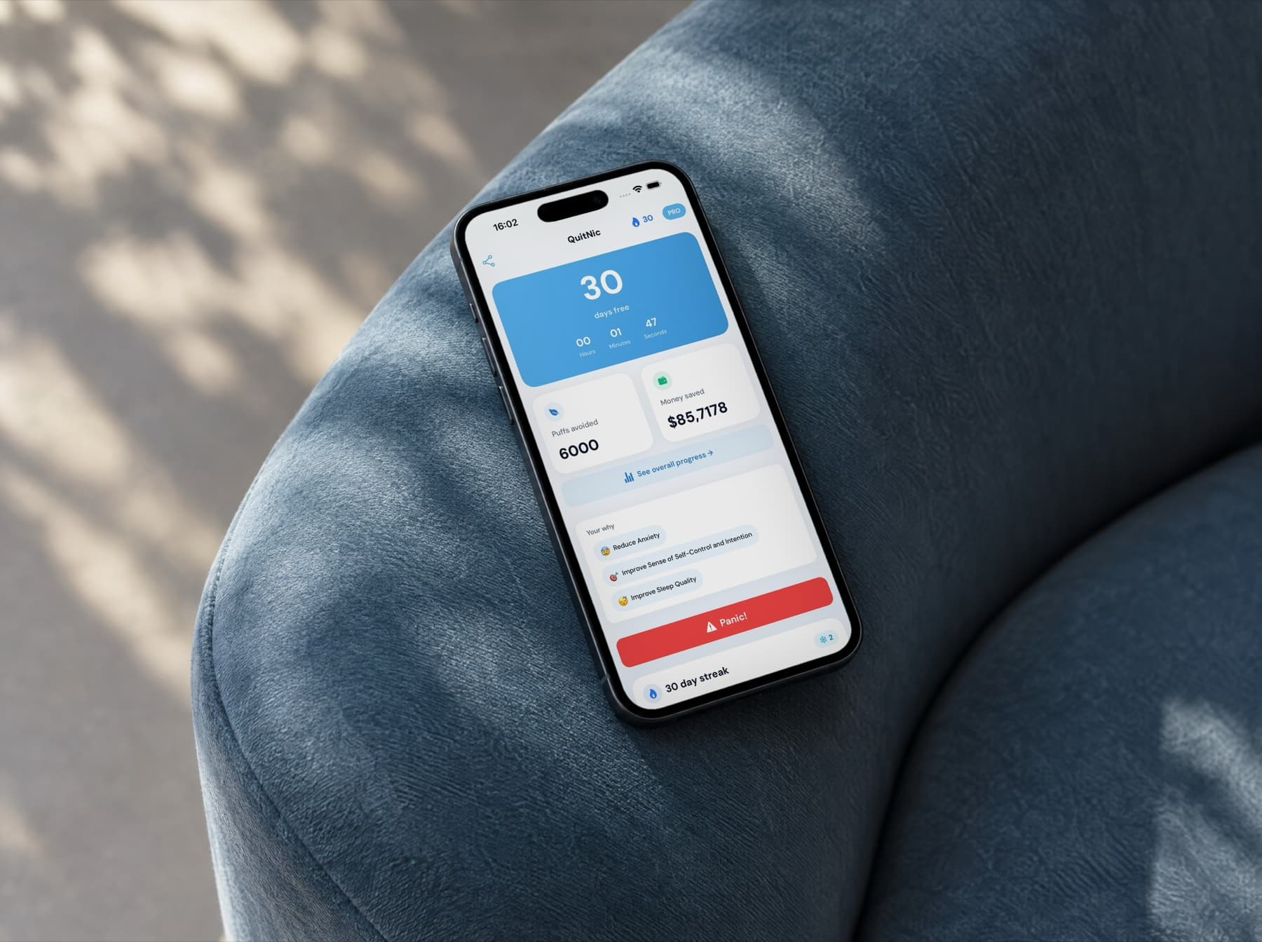 An iPhone on the arm of a blue armchair in morning light. The screen shows the QuitNic dashboard: 30 days nicotine-free, with puffs avoided and money saved.