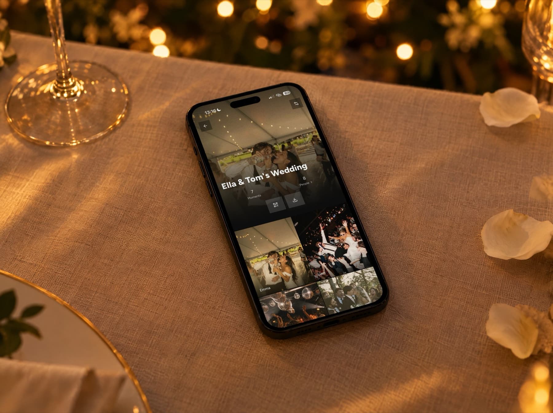 An iPhone on a linen-covered wedding table in evening light. The screen shows the Unveil app: a shared wedding album with photos taken by the guests.